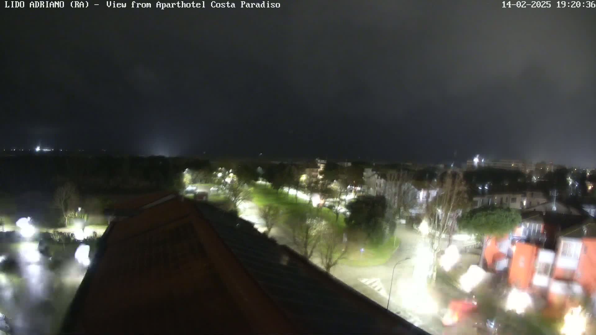 A nighttime, overcast view of a town with roads, trees, and buildings illuminated by streetlights, possibly during a light rain or mist.