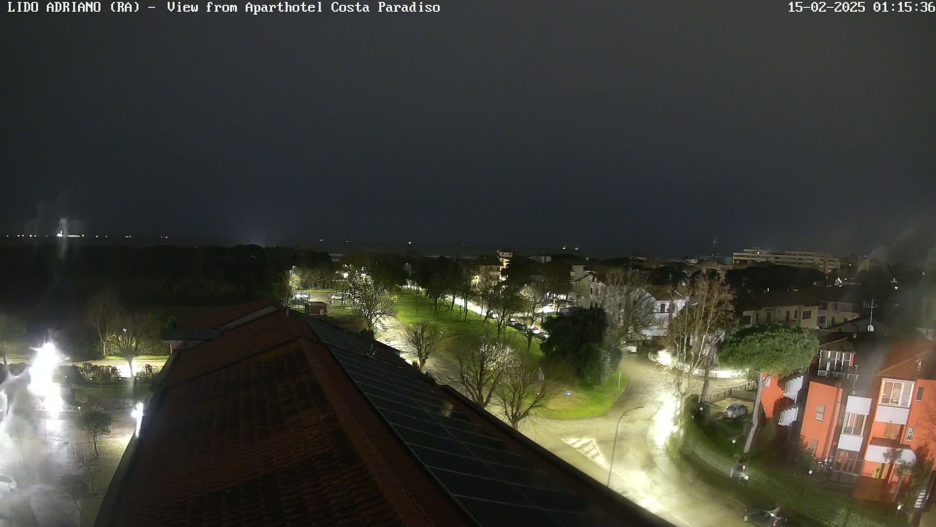 A nighttime view of a town under a dark, overcast sky showing rain-slicked streets, illuminated buildings, and trees.