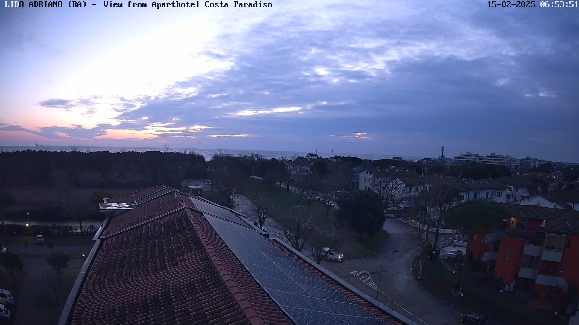 A sunrise over a calm sea is visible in the distance behind a residential area, viewed from a rooftop with solar panels.