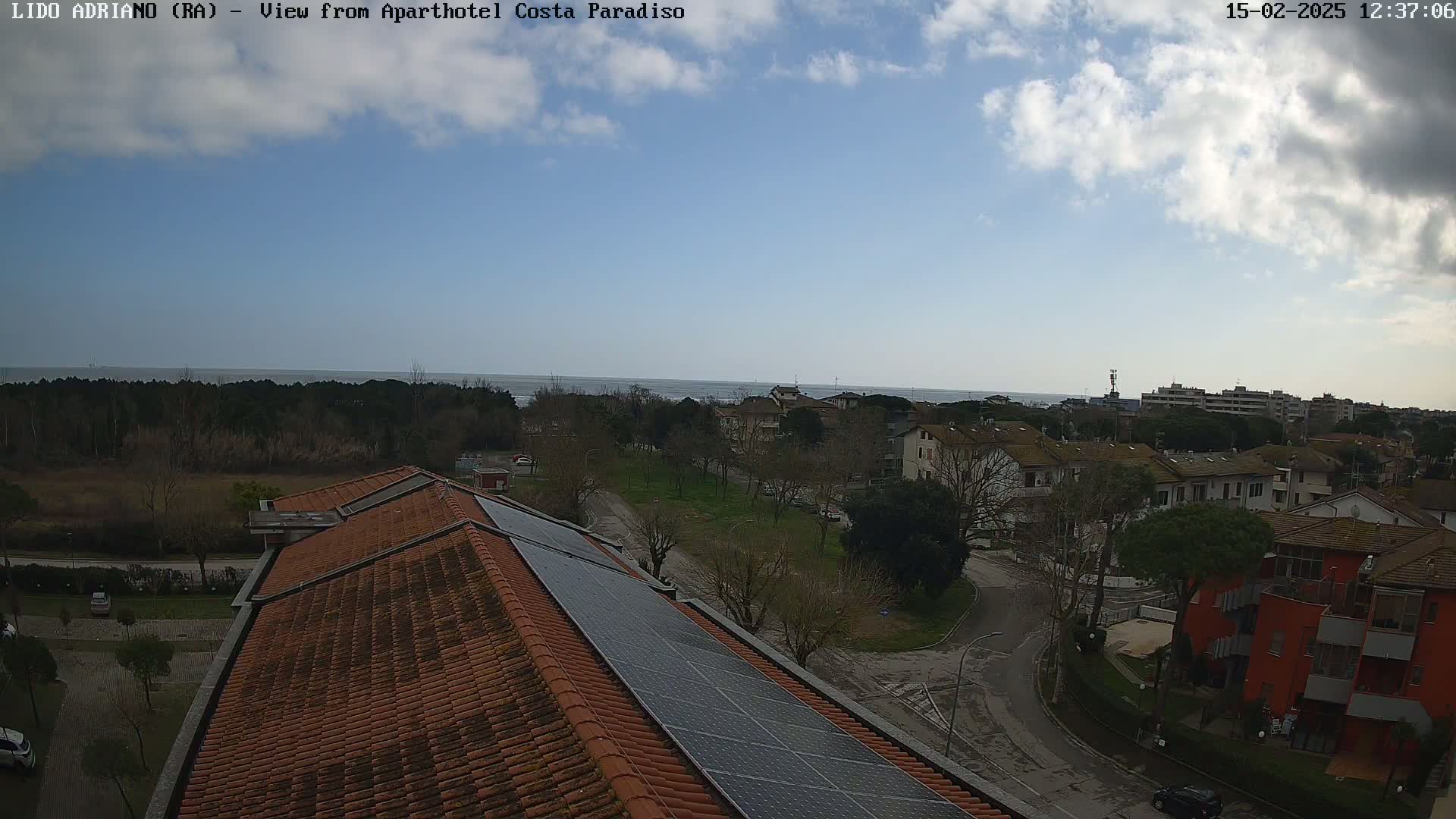 A partly cloudy day reveals a view of a coastal town with a calm sea in the distance, seen from a rooftop with solar panels.