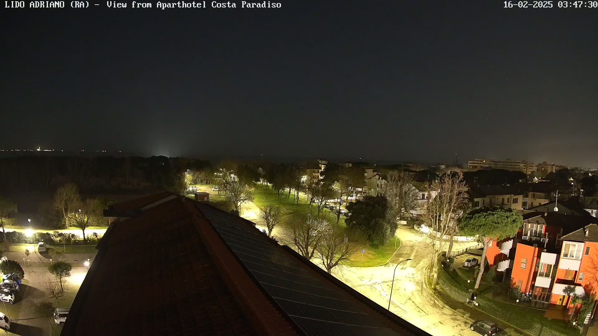 A nighttime view of a residential area near a coastline, illuminated by streetlights under a dark, clear sky.