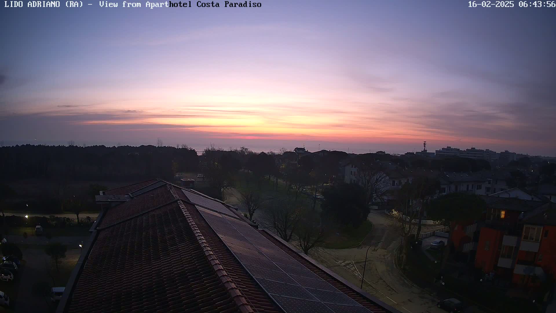 A sunrise over a coastal town, viewed from a rooftop with solar panels, under a mostly clear sky.