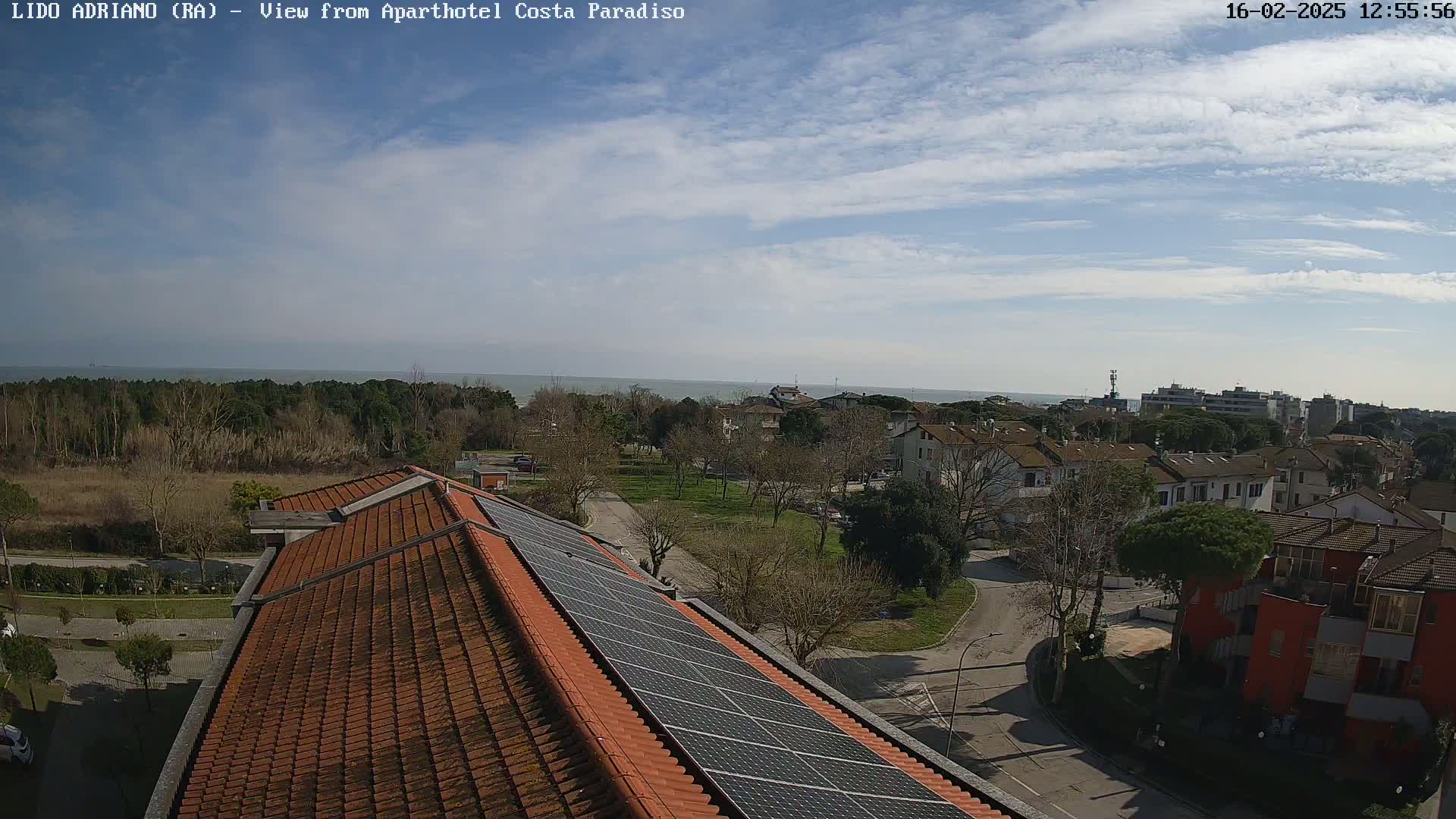 A partly cloudy sky overlooks a coastal town with red-tiled roofs and solar panels on a building, featuring a calm sea in the distance.