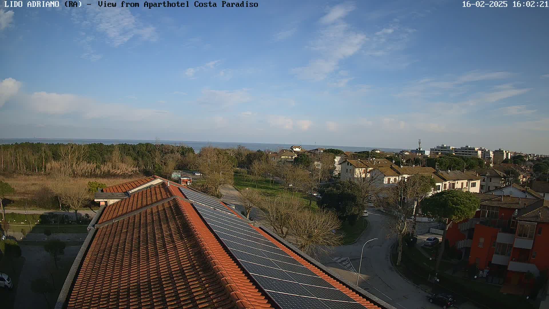 A mostly sunny day overlooks a coastal town with red-tiled roofs and solar panels on a building in the foreground, with a calm ocean visible in the distance.
