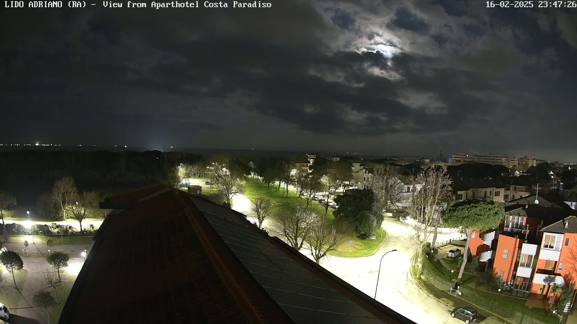 A nighttime view of a coastal town under a cloudy moonlit sky shows a roadway with streetlights illuminating buildings and trees, with a body of water visible in the distance.