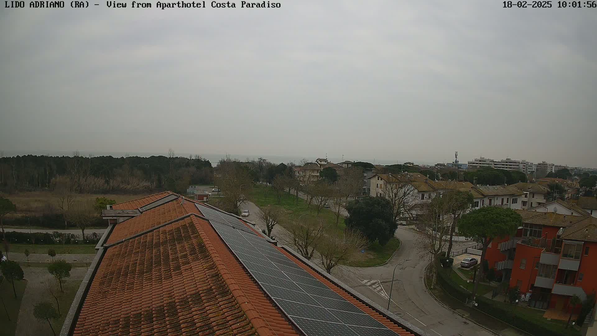 An overcast day reveals a coastal town's rooftops, a tree-lined park, and a calm sea in the distance, all viewed from a building with solar panels on its roof.