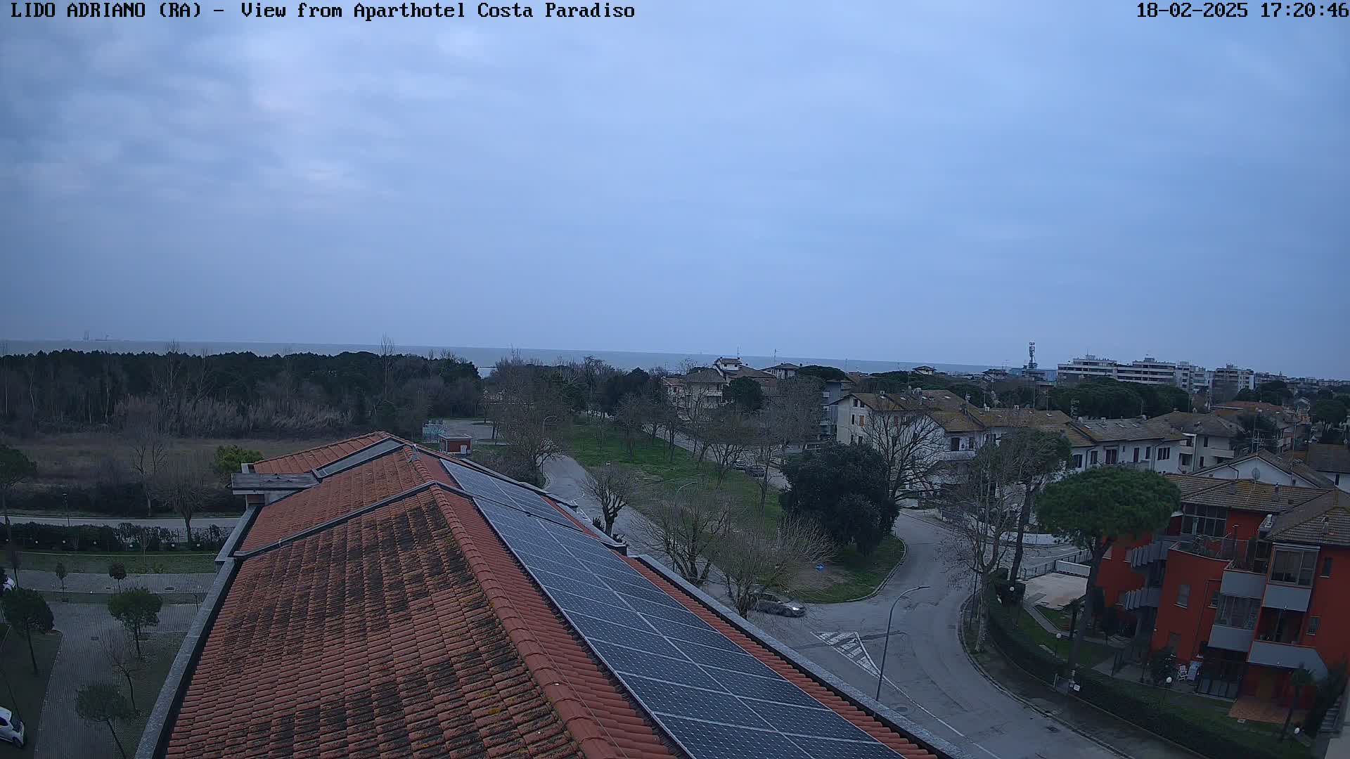 A mostly cloudy day overlooks a coastal town with red-tiled roofs and solar panels on a building, a calm sea visible in the distance.