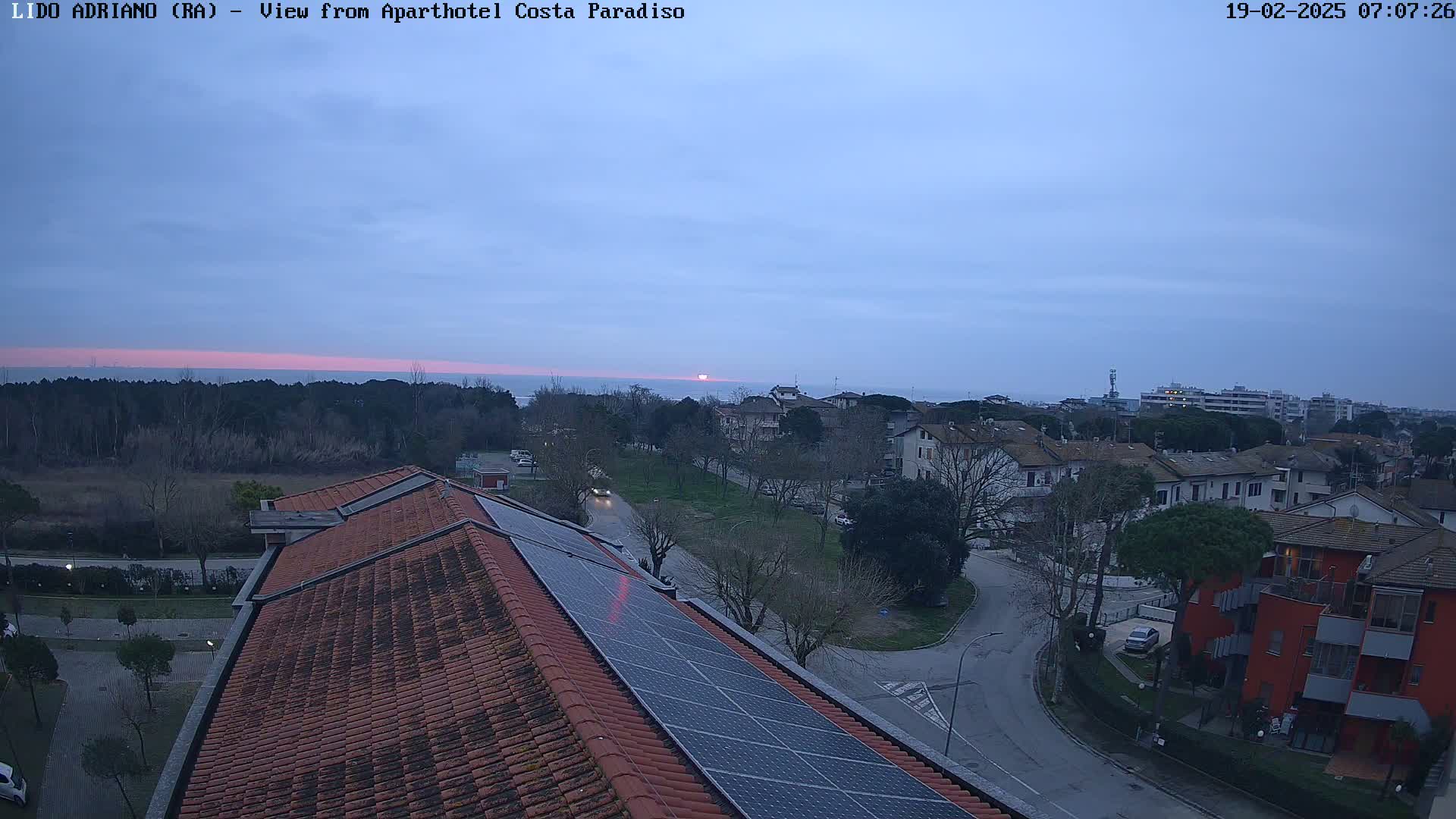 A sunrise over a calm sea is visible from a rooftop with solar panels overlooking a residential area.