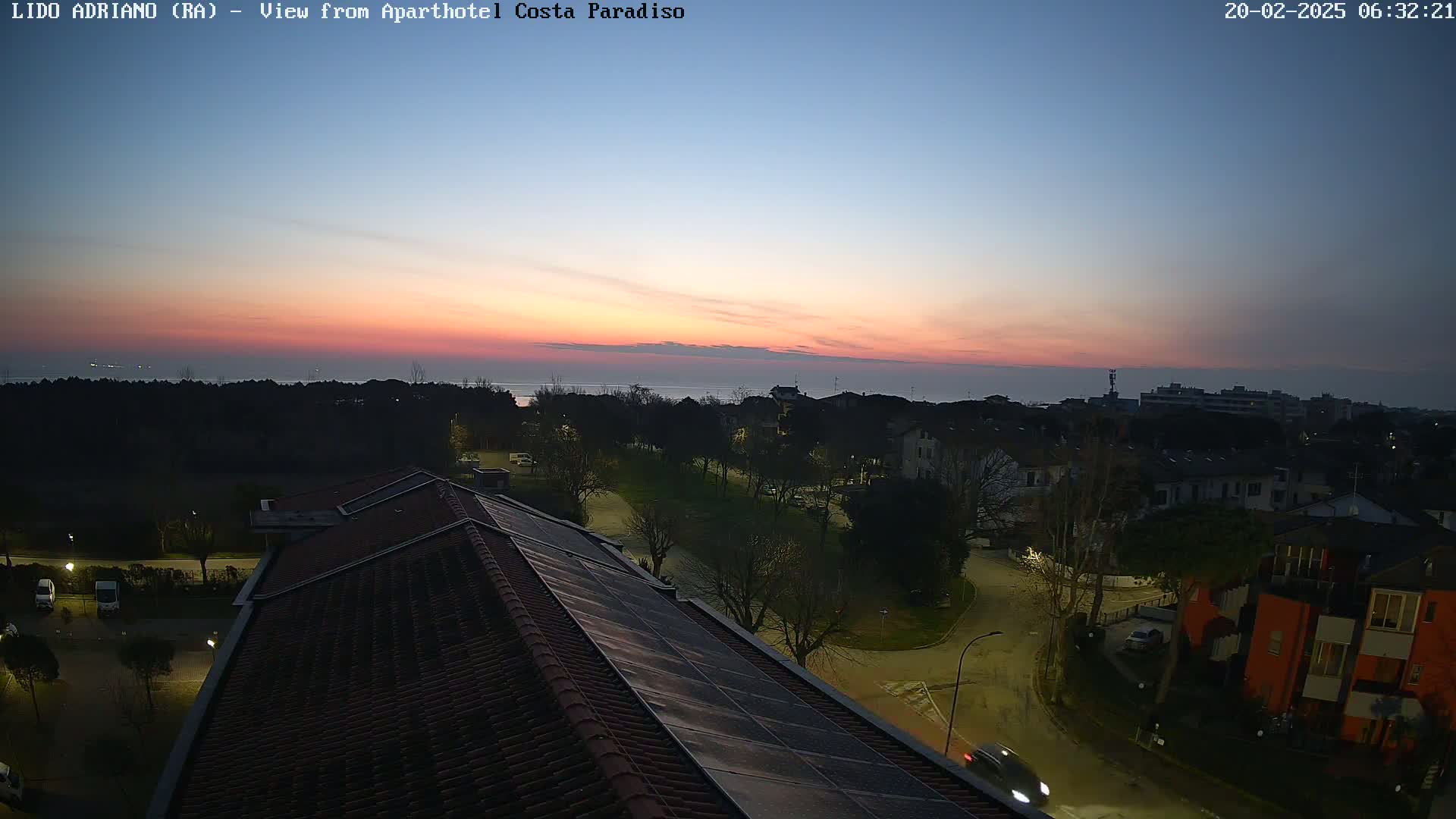 A seaside town at sunrise, viewed from a rooftop with solar panels, showing a calm sea and residential streets below.