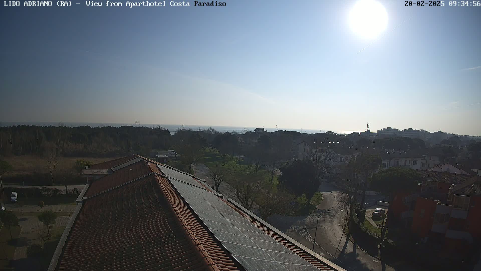 A sunny day reveals a view of a seaside town from a rooftop with solar panels, showing a calm ocean and residential buildings.
