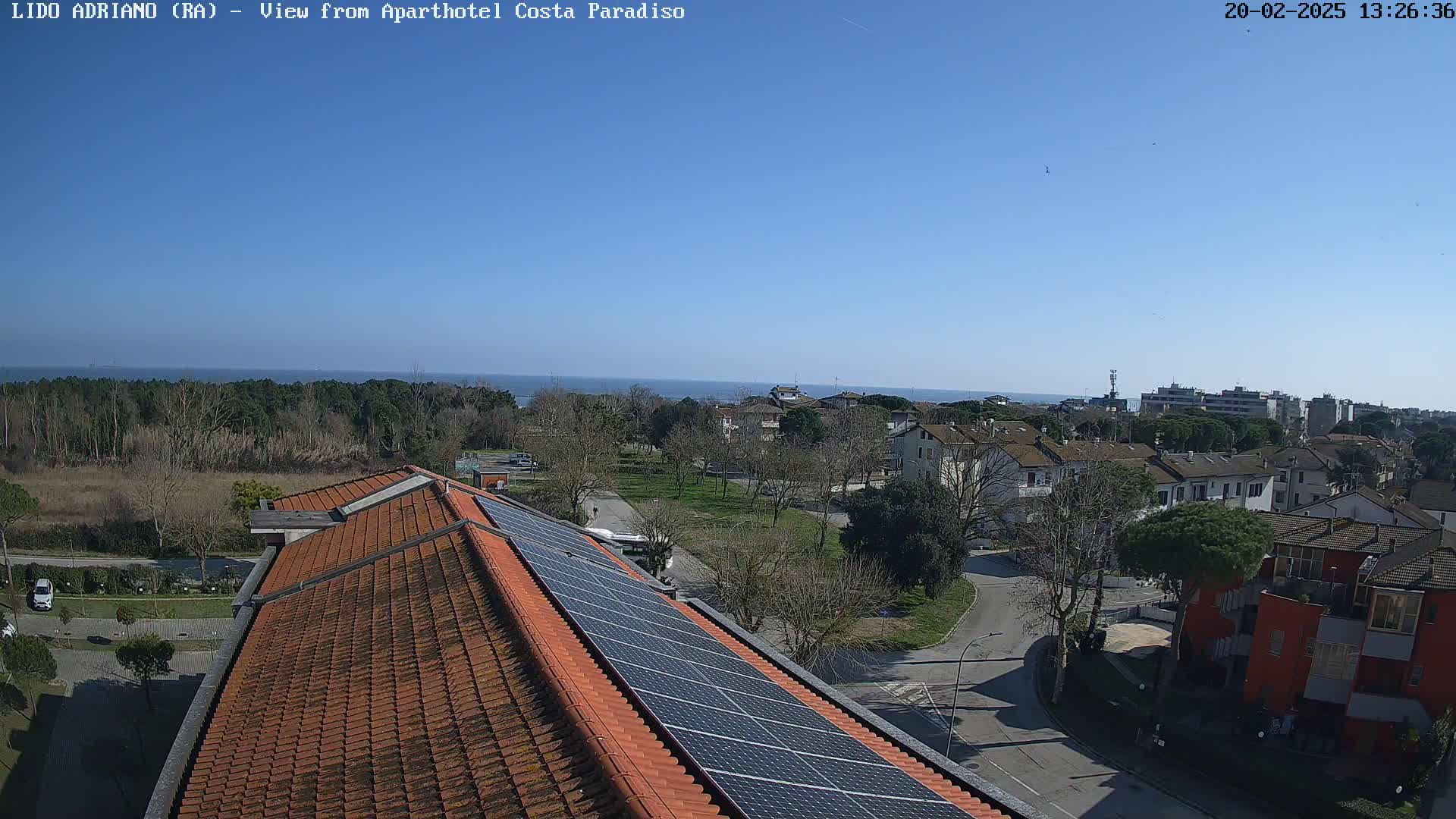 A clear, sunny day overlooks a coastal town with red-tiled rooftops, a park, and a sea view, where a building with solar panels is prominently featured in the foreground.