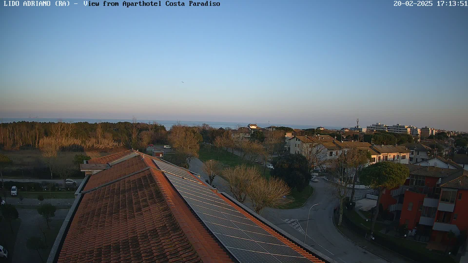 A clear, late afternoon view of a coastal town shows a residential area with a calm sea in the background, partially visible behind a line of trees.