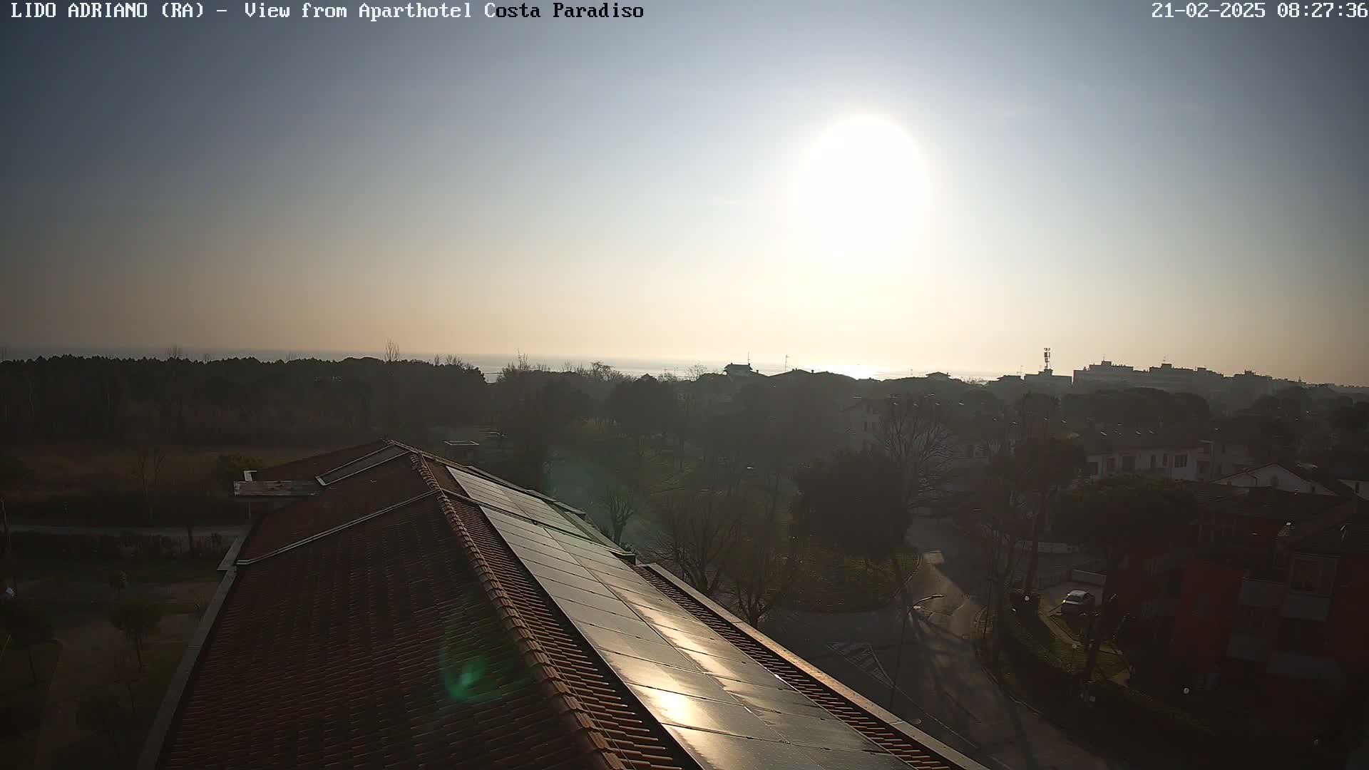 A view from a rooftop shows a hazy sunrise over a calm sea and a residential area with trees.