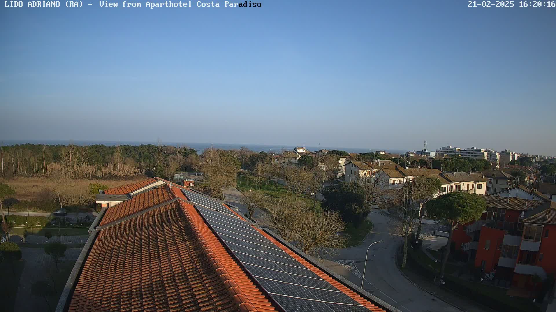 An elevated view shows a tiled roof with solar panels overlooking a residential area near a calm ocean under a clear blue sky.