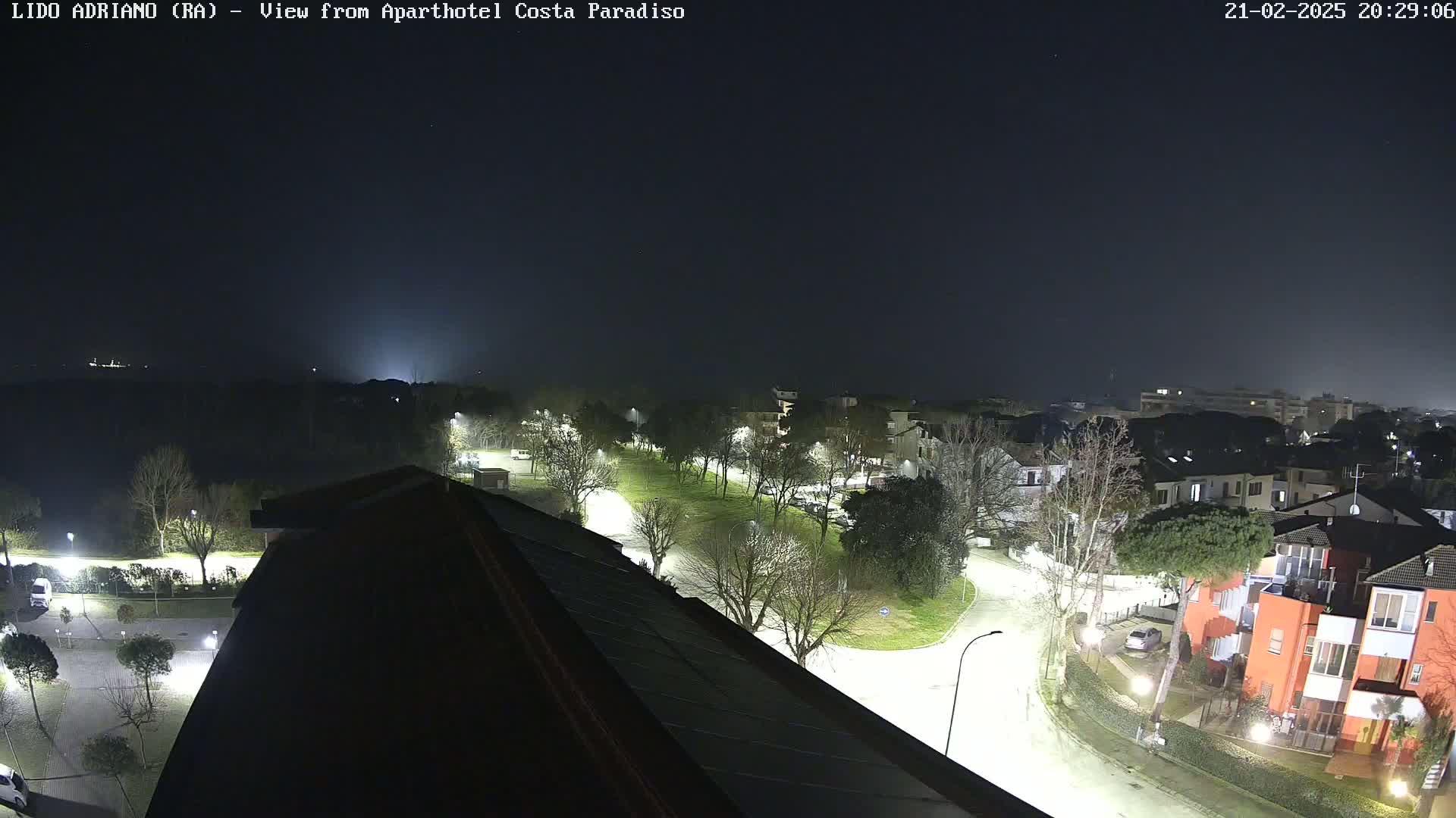 A nighttime, slightly hazy view from a rooftop shows a residential area with illuminated streets and buildings, and a small park.