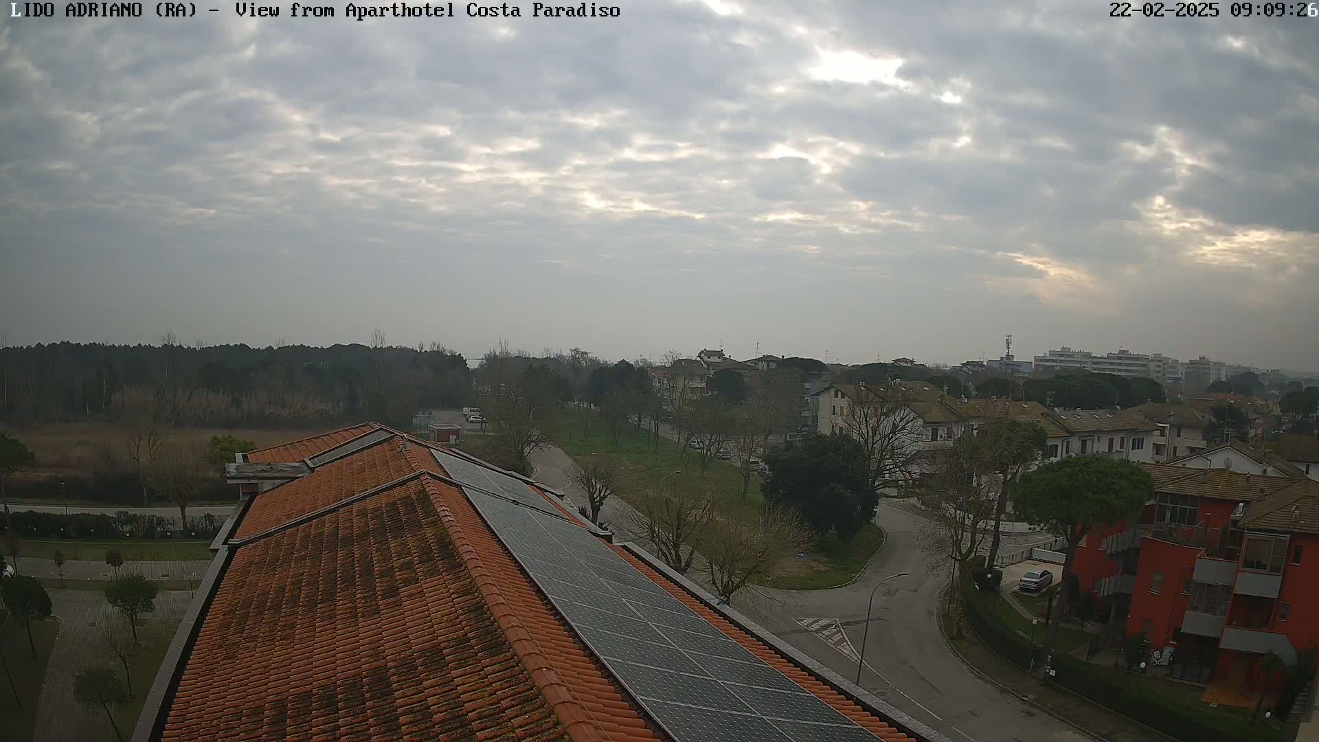 A cloudy day reveals a view of a residential area with a mix of buildings, trees, and a road, seen from a rooftop with solar panels.