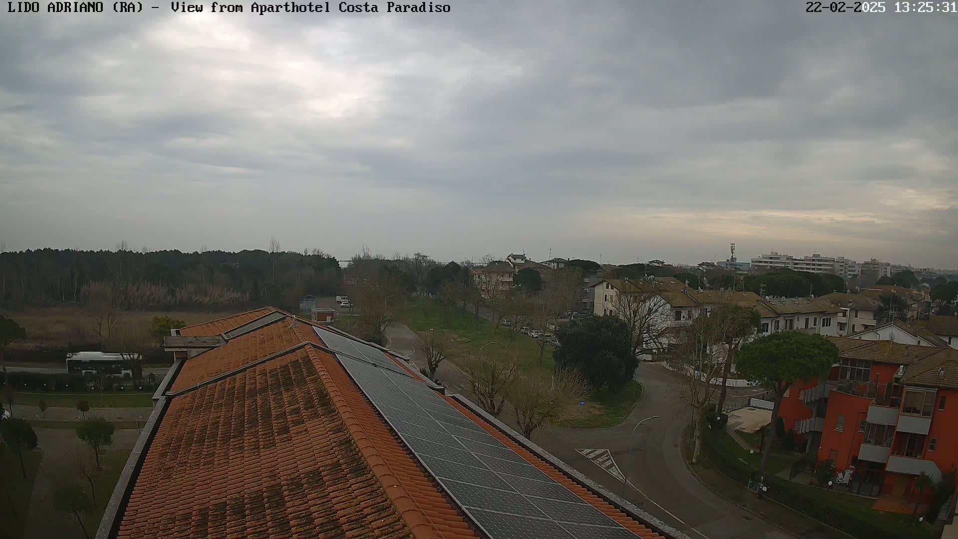 A view from a rooftop with solar panels shows a cloudy day over a residential area with trees, roads, and a bus.