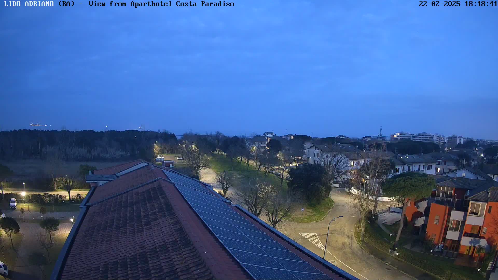 A rooftop view at twilight shows a residential area with roads and trees, under a clear, dark blue sky.