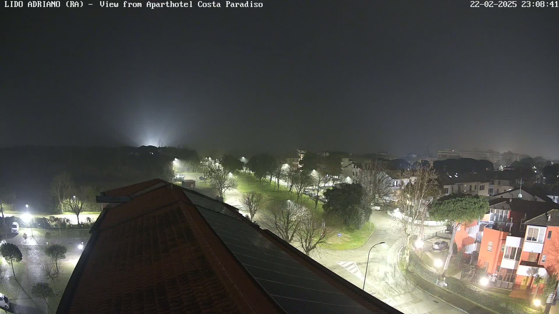 A nighttime view of a residential area under a foggy sky, showing wet roads and illuminated buildings.