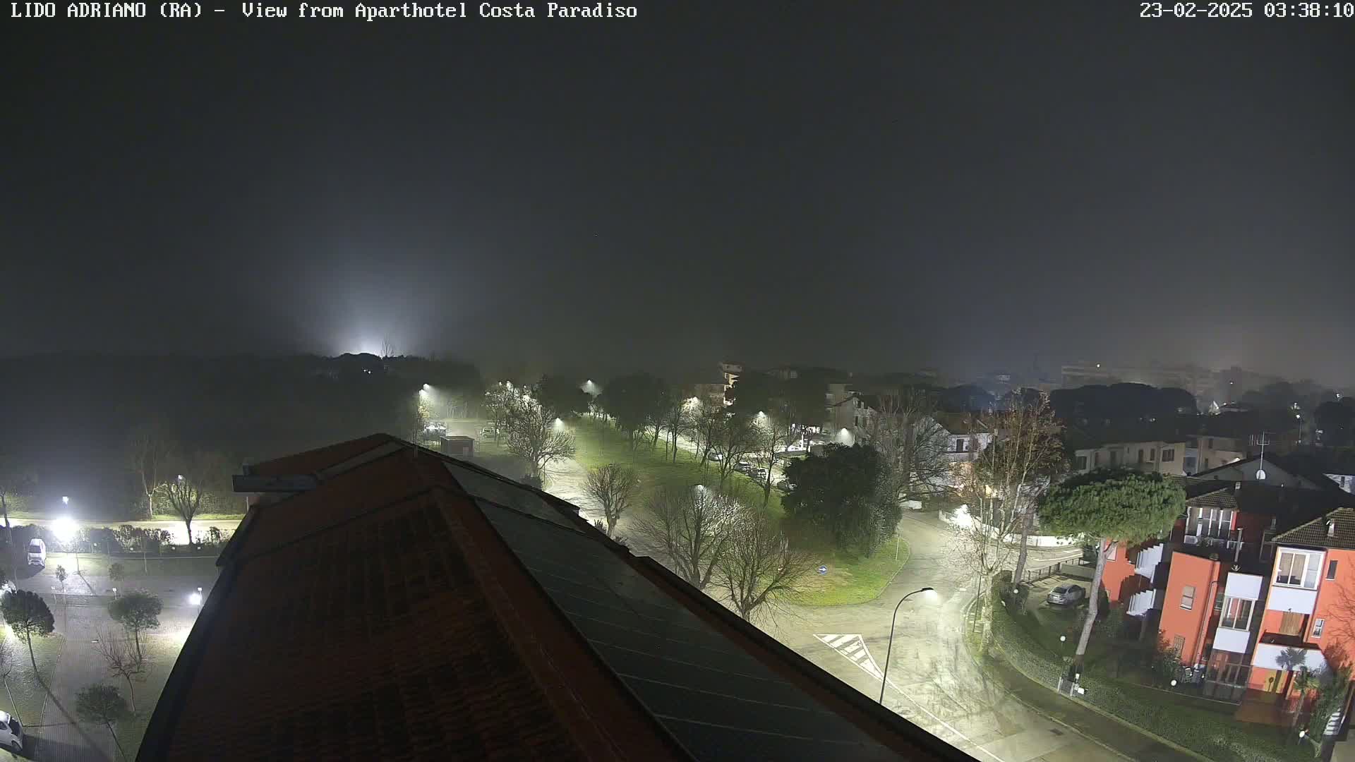 A nighttime, foggy view from a rooftop shows a residential area with streetlights illuminating roads and buildings.