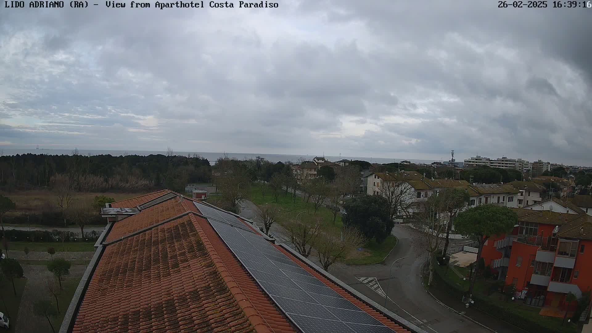 An overcast sky overlooks a coastal town with a red-tiled building with solar panels on the roof, a calm sea in the distance.