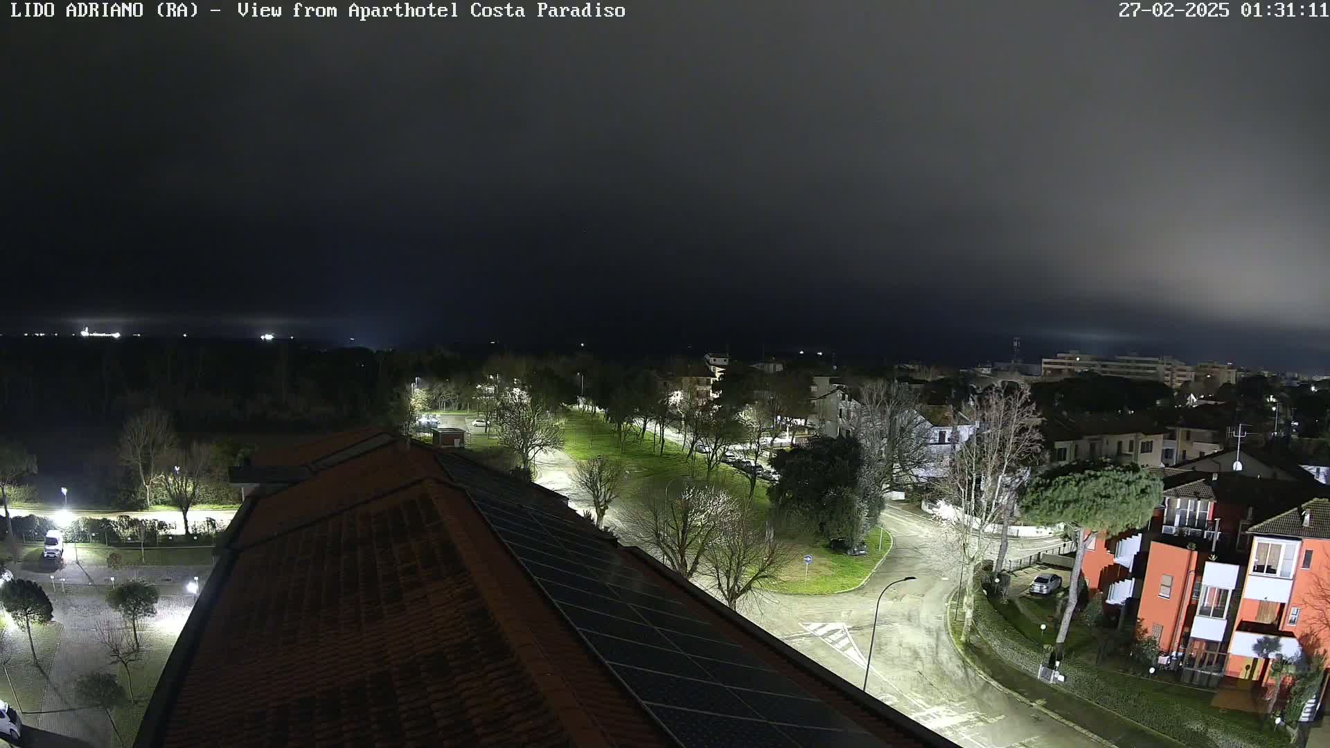 A nighttime, foggy view of a residential area with streetlights illuminating roads and buildings, seen from a rooftop with solar panels.
