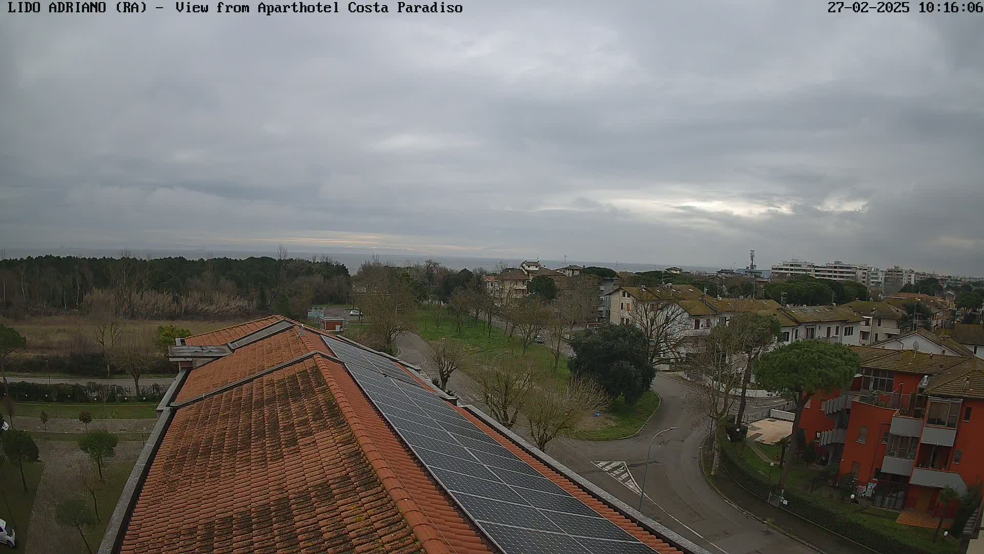 An overcast sky overlooks a coastal town with low-rise buildings and a large solar panel array on a red-tiled roof.