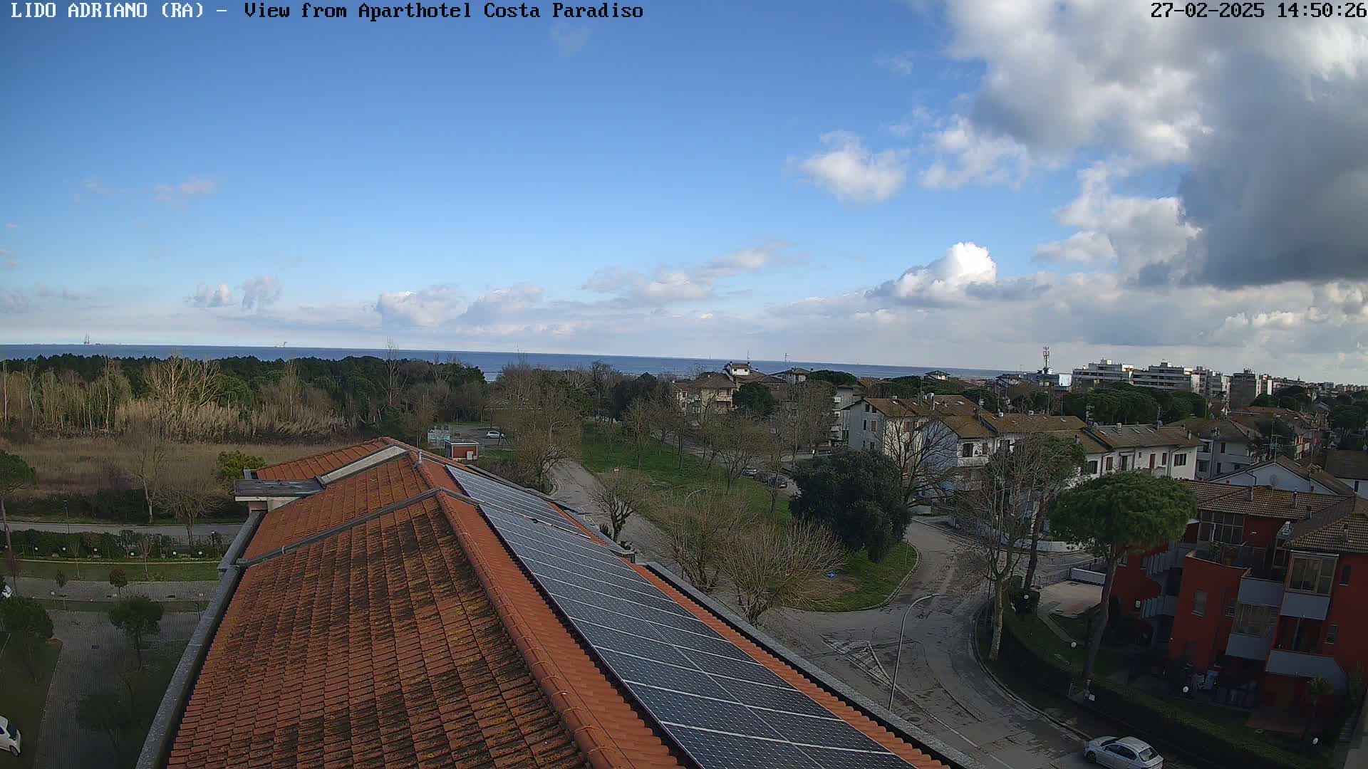 A partly cloudy day overlooks a coastal town with a building featuring solar panels on its roof, and a calm sea in the distance.