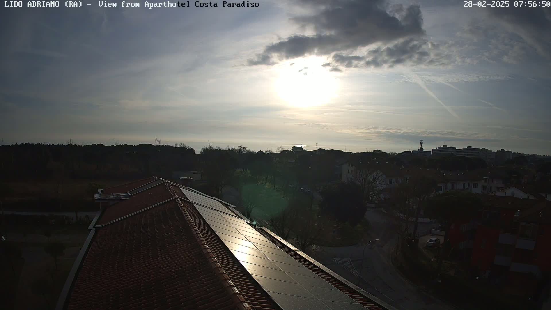 A partly cloudy sunrise over a residential area with a view of the sea in the distance, seen from a rooftop with solar panels.