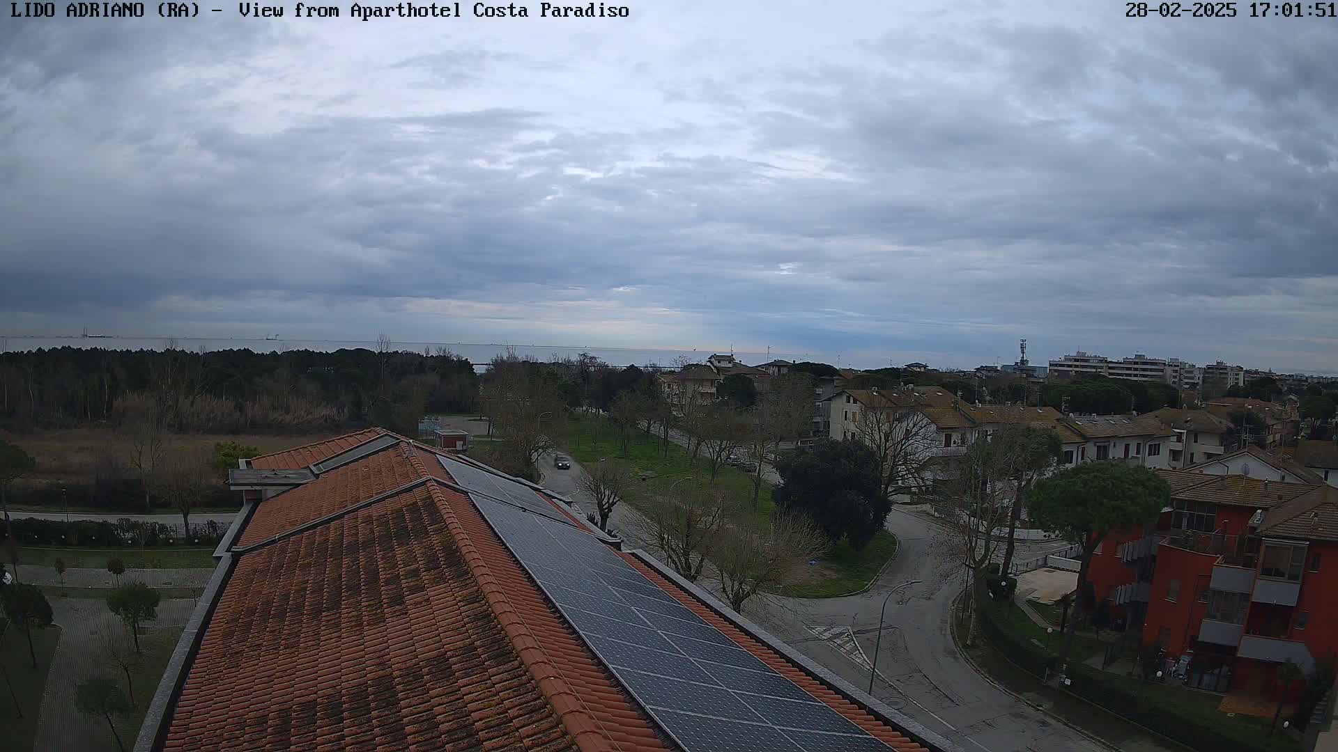 A view from a rooftop with solar panels shows a mostly cloudy sky over a residential area near a calm body of water.