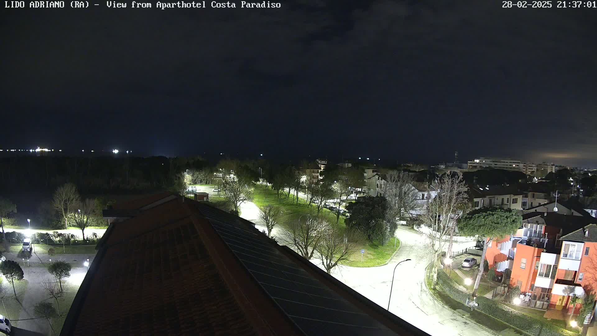 A nighttime aerial view shows a residential area with illuminated streets and buildings, under a dark, cloudy sky.