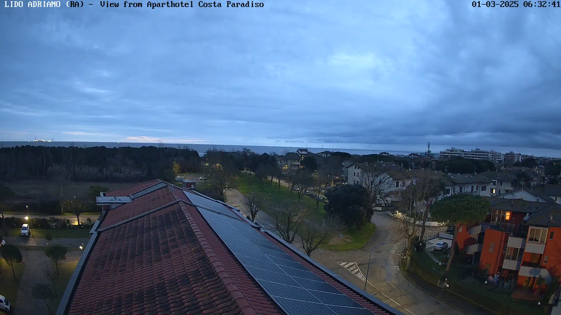 A view from a rooftop with solar panels shows a coastal town under a cloudy, twilight sky.