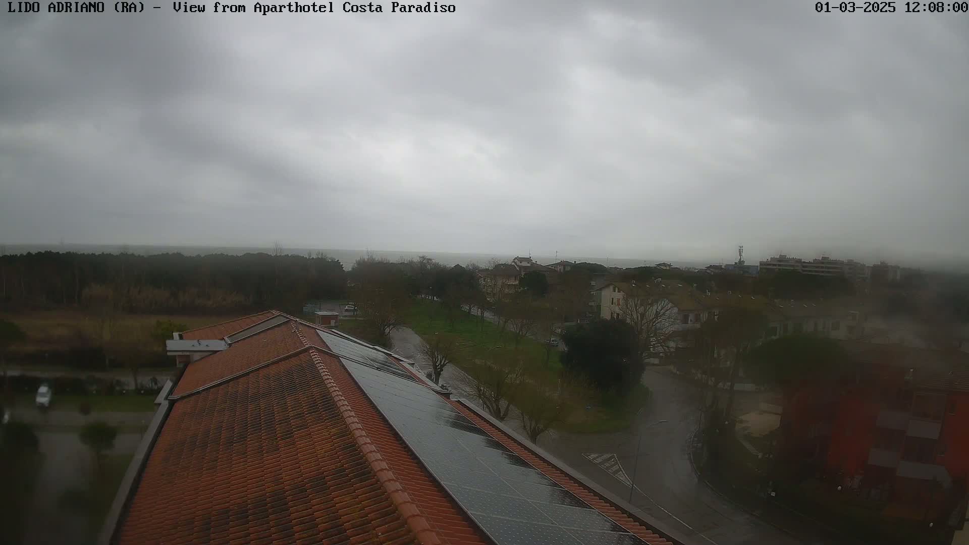 A view from a rooftop with a red tile roof and solar panels shows a rain-soaked residential area extending to a calm sea under a cloudy sky.