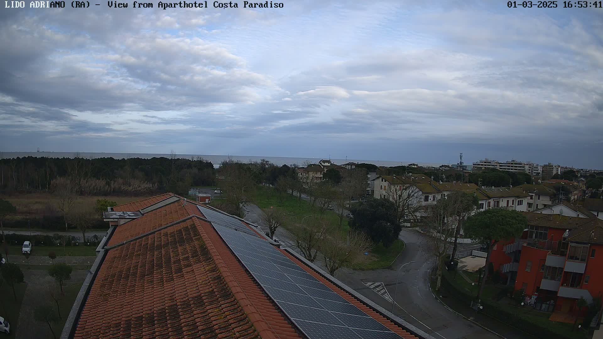 A view of a coastal town under a cloudy sky, showing rooftops with solar panels and a calm sea in the distance.