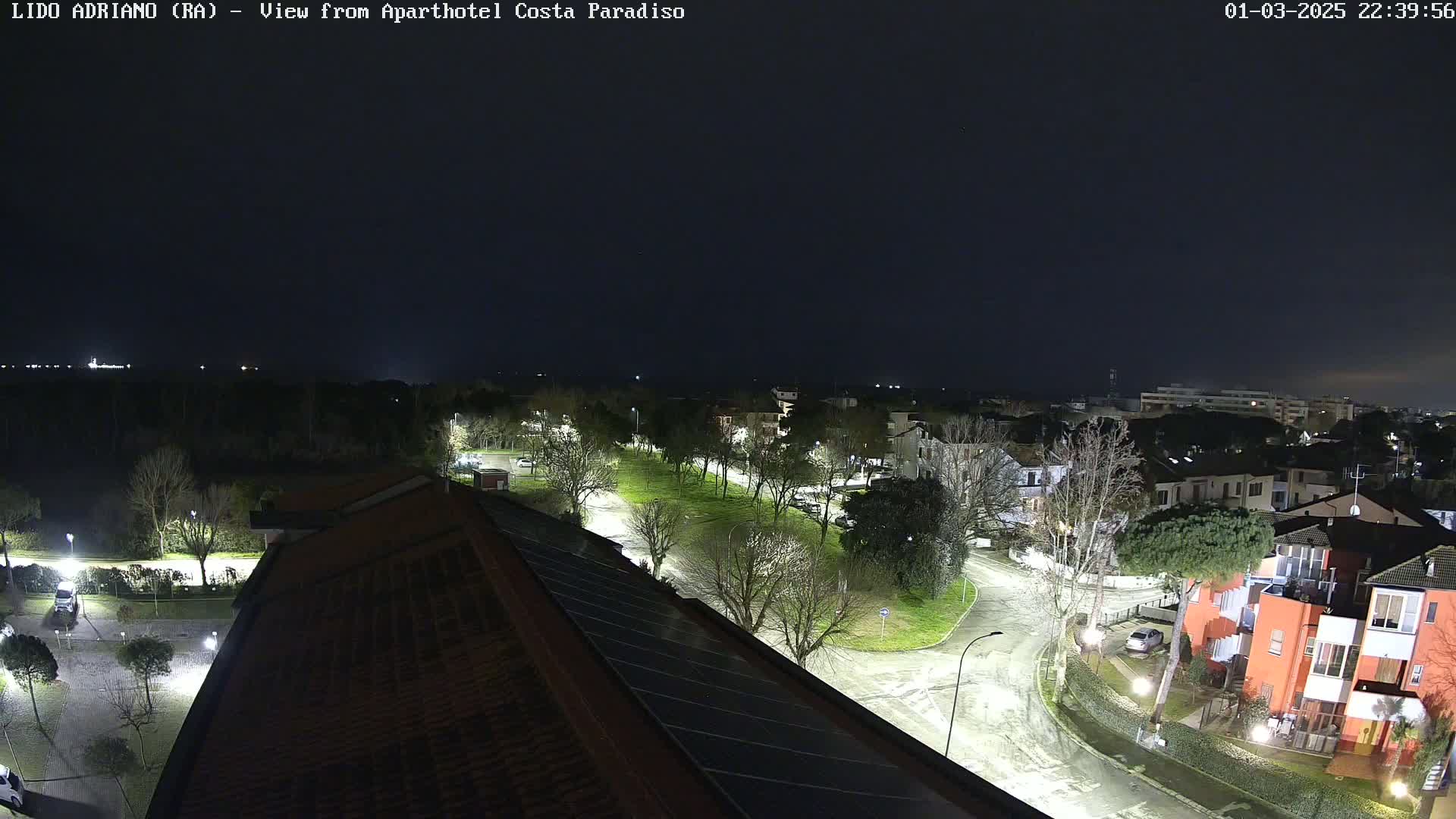 A nighttime aerial view of a residential area with tree-lined streets and buildings, illuminated by streetlights under a dark sky.