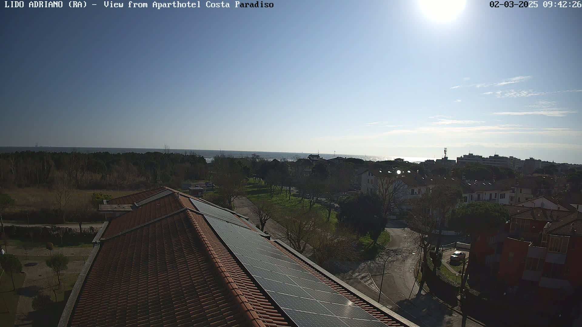 A sunny day overlooking a coastal town with residential buildings and a rooftop equipped with solar panels, featuring a calm sea in the background.