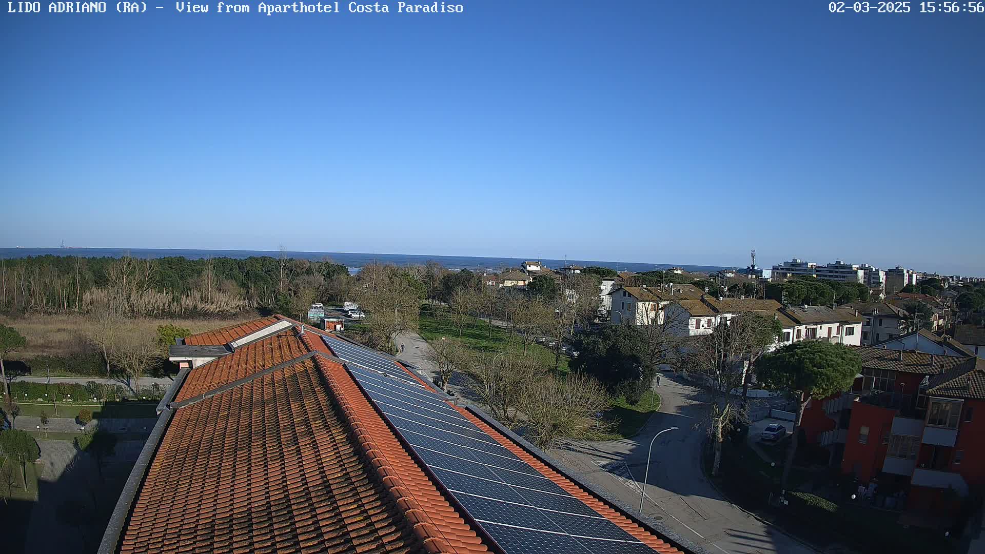 A clear, sunny day overlooking a coastal town with a tiled roof and solar panels in the foreground, and a calm sea visible in the distance.