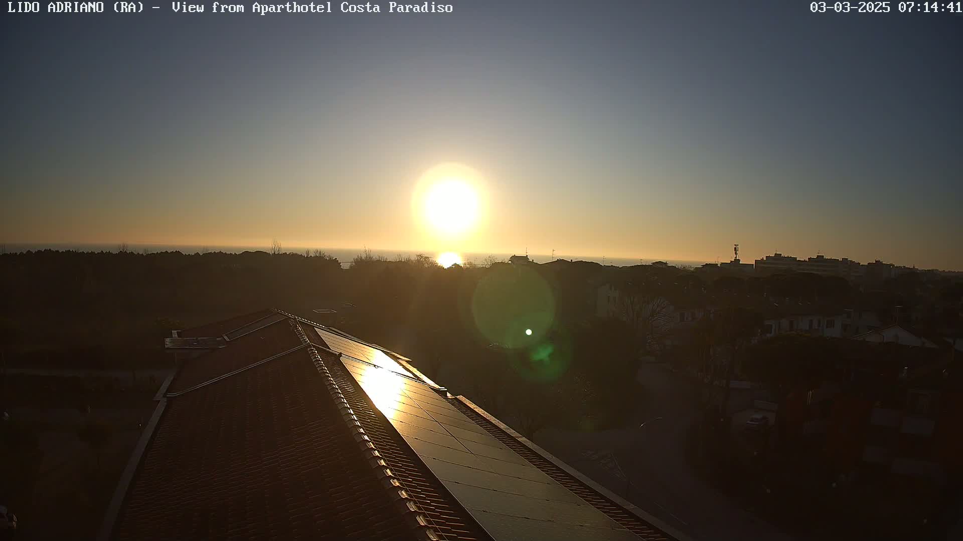 A sunrise over a calm ocean is seen from a rooftop with solar panels, overlooking a residential area.