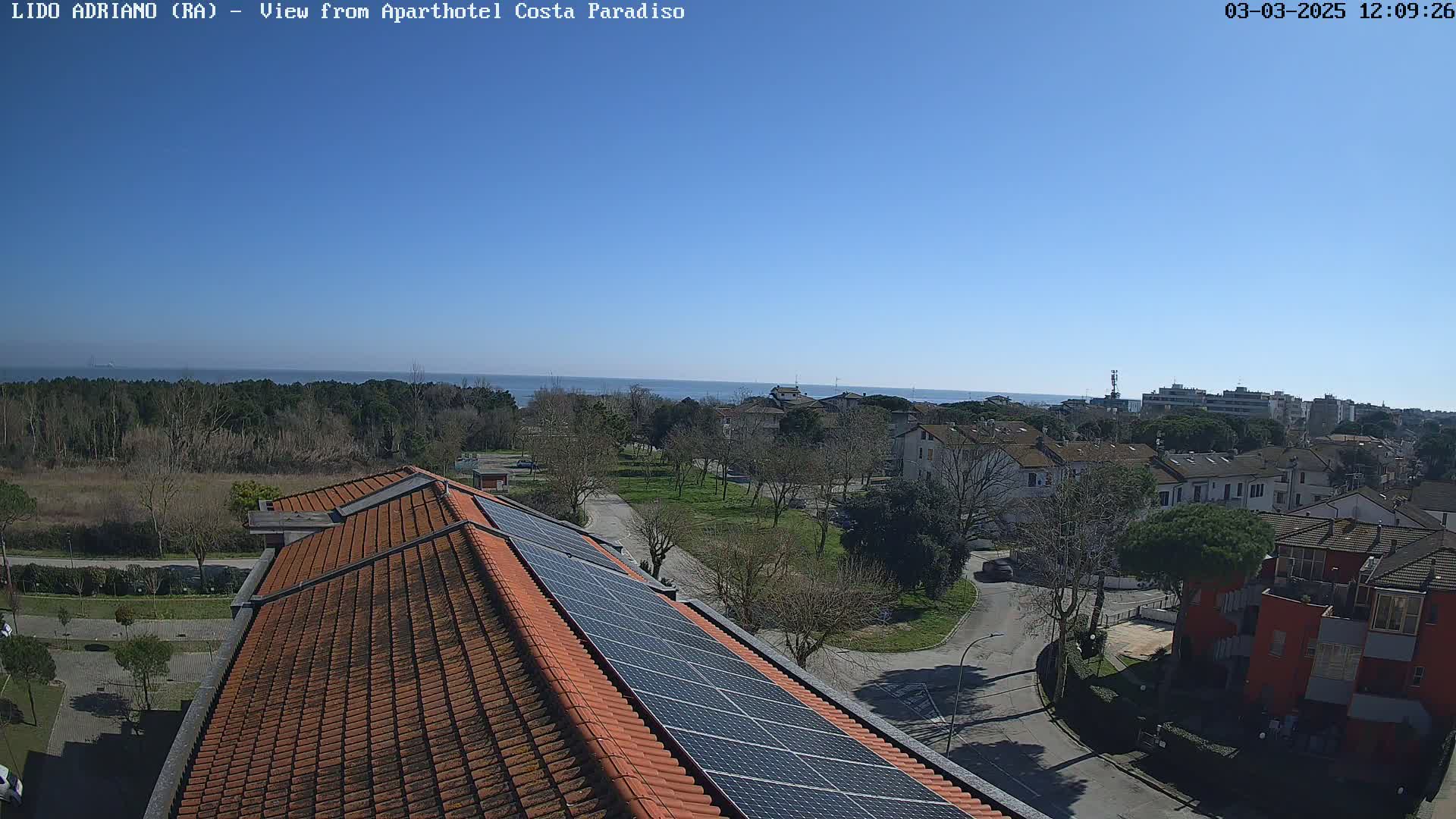 A clear, sunny day overlooking a coastal town with residential buildings, trees, and a rooftop equipped with solar panels; a calm sea is visible in the distance.