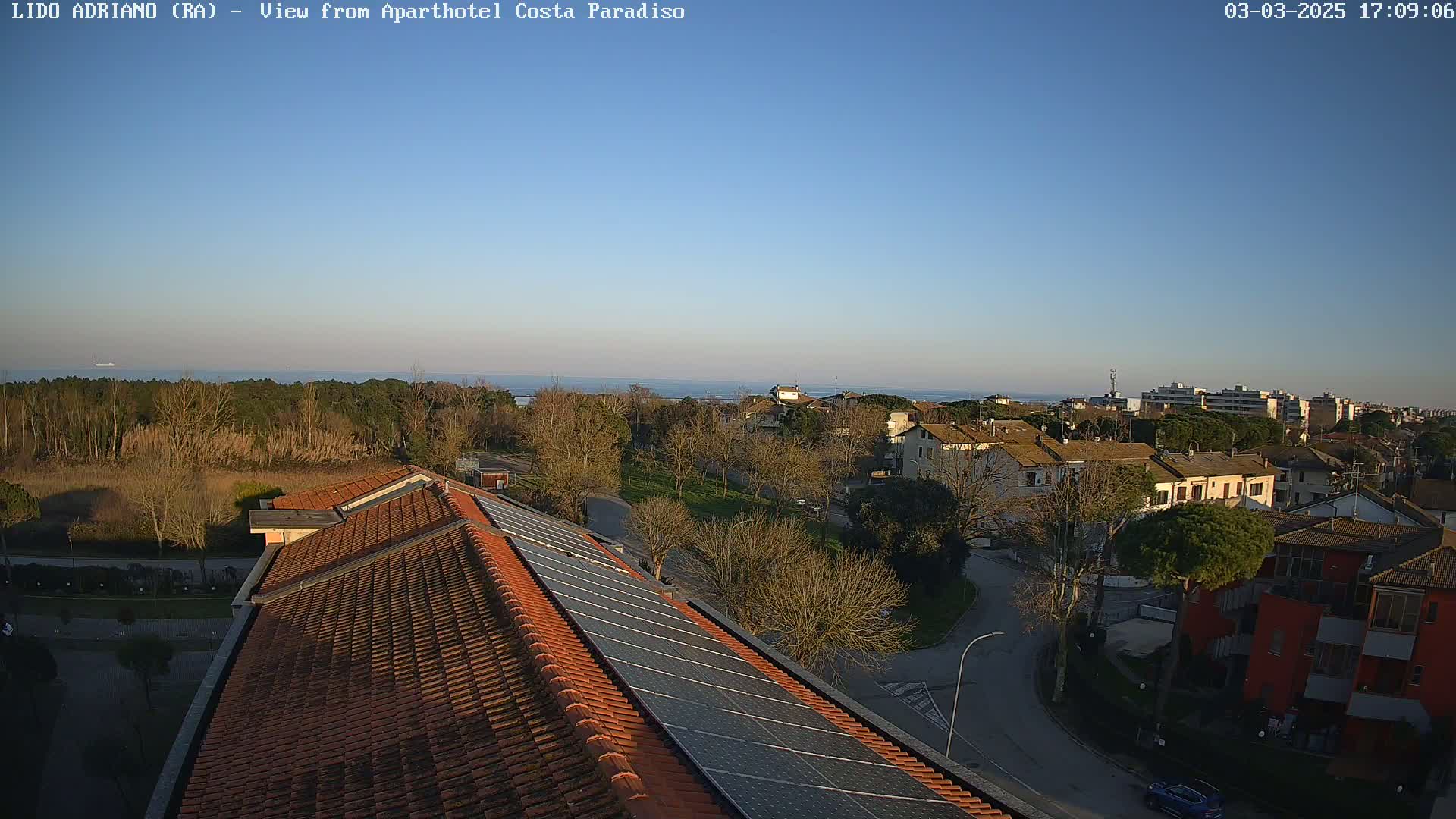 A clear, sunny day overlooks a coastal town with residential buildings, trees, and a calm ocean in the distance, viewed from a rooftop with solar panels.