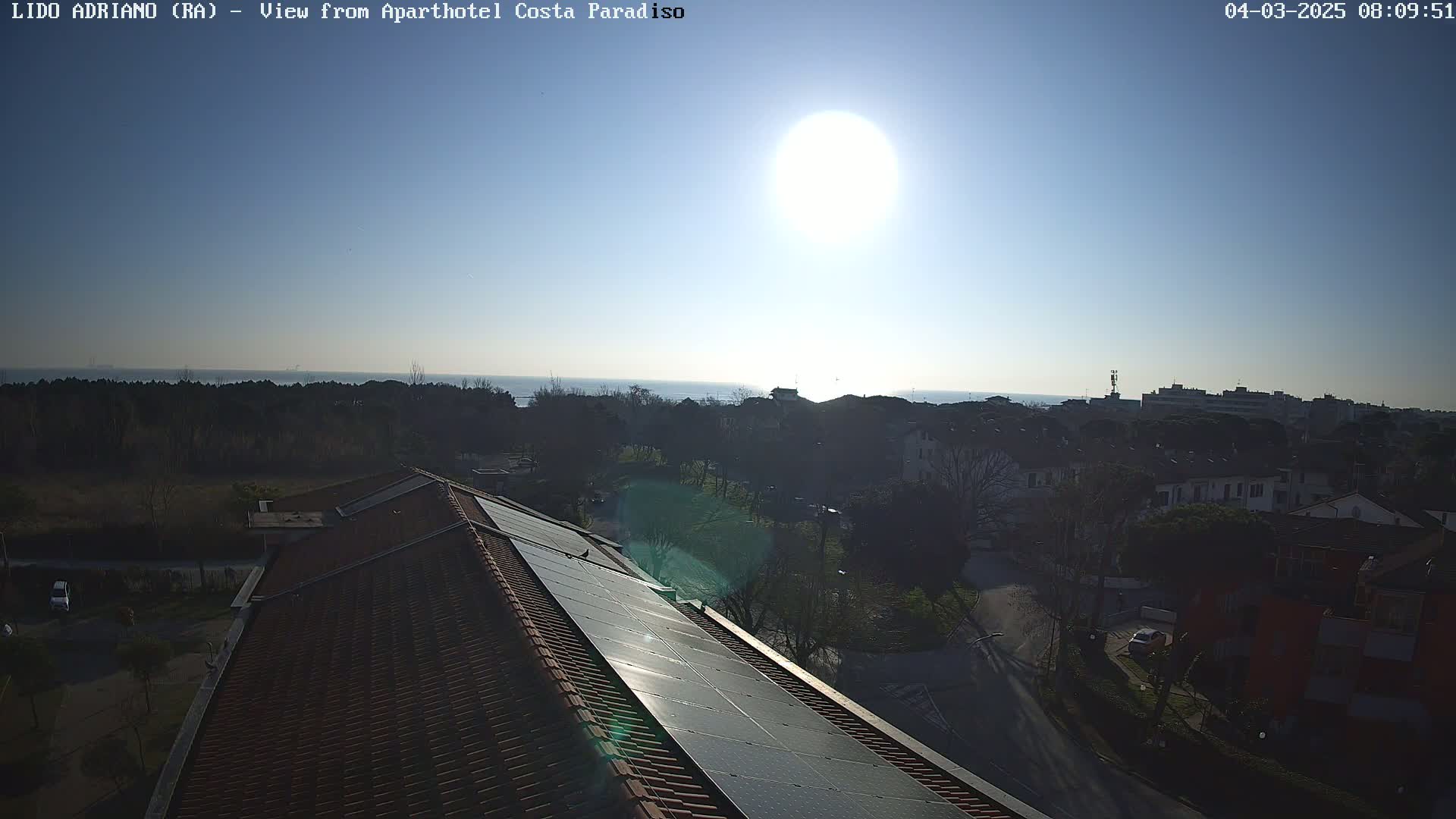 A rooftop with solar panels overlooks a sunny residential area and a calm sea in the distance.