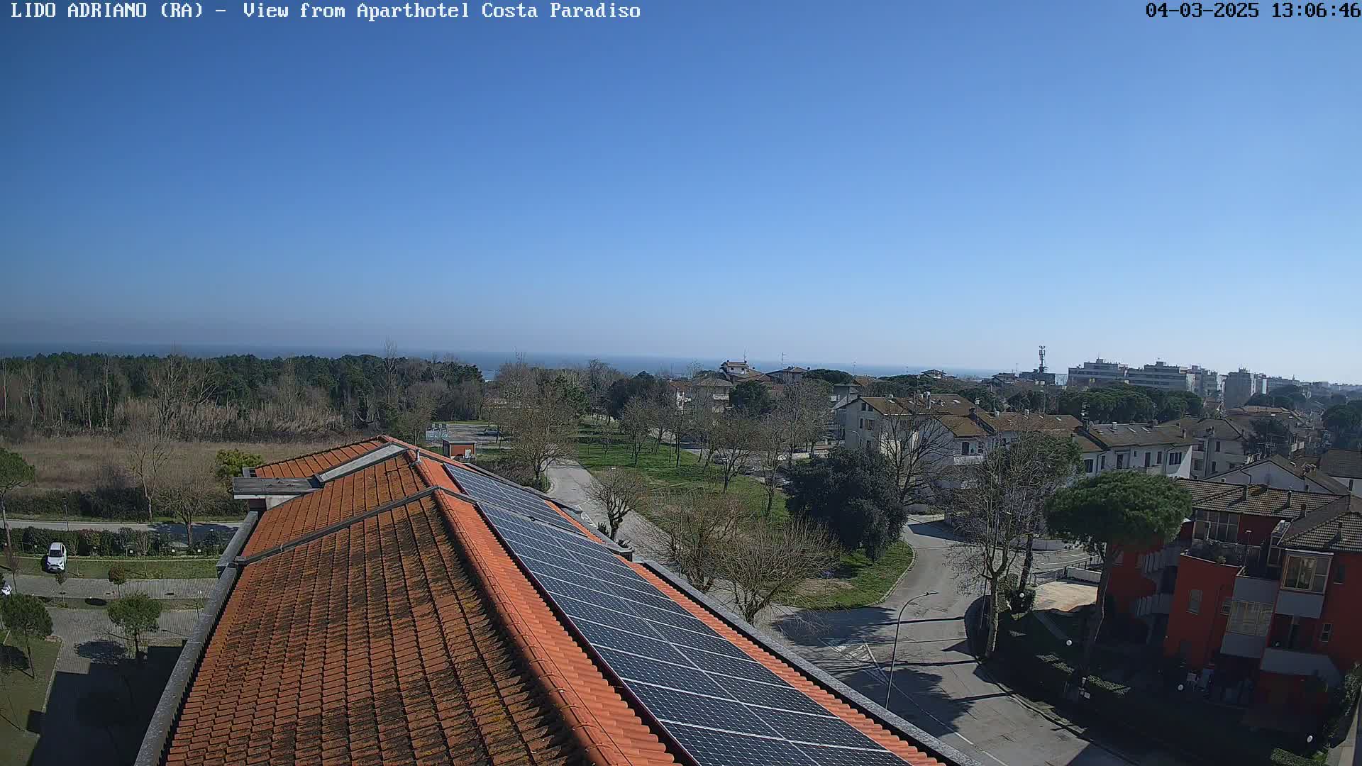 A clear, sunny day overlooks a residential area with a sea view, featuring a building rooftop with solar panels.