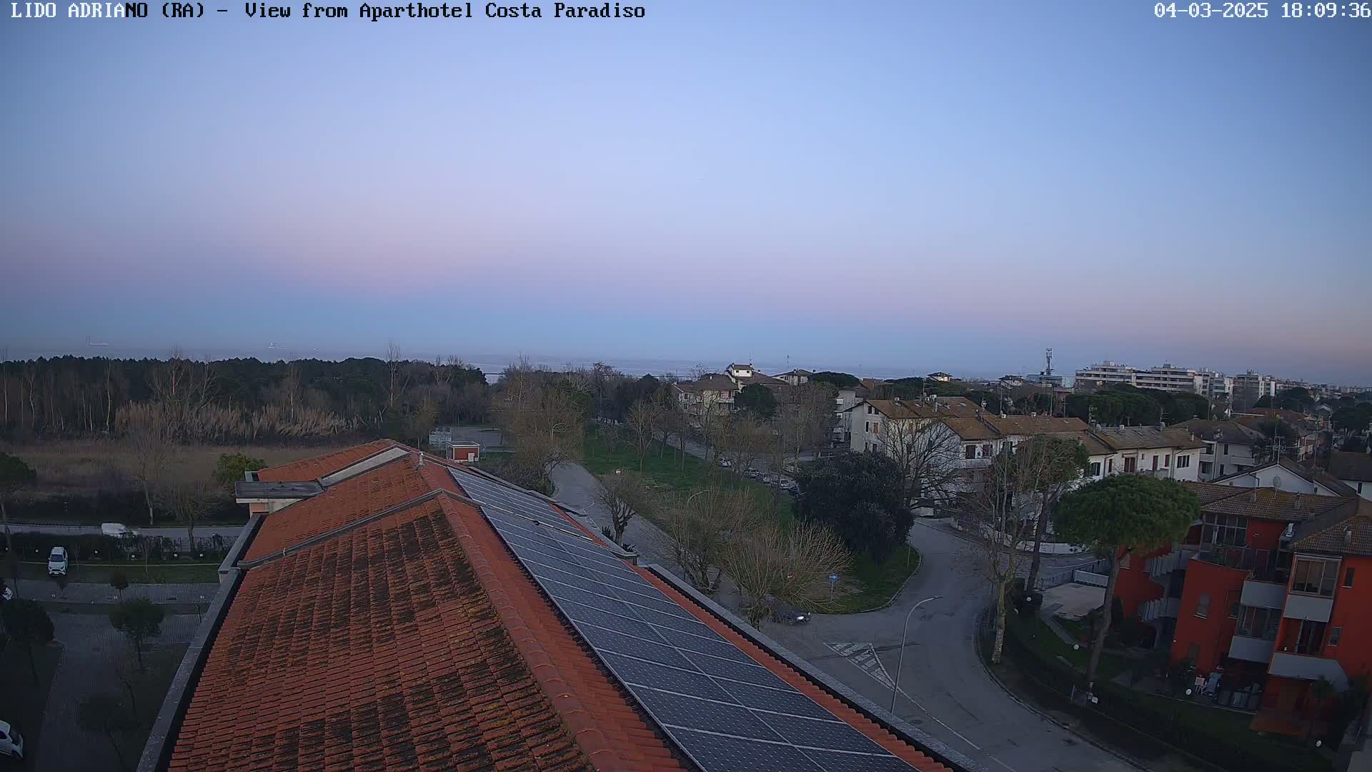 A high-angle view shows a residential area at dusk, with a calm body of water visible in the distance under a clear, pale-purple sky.