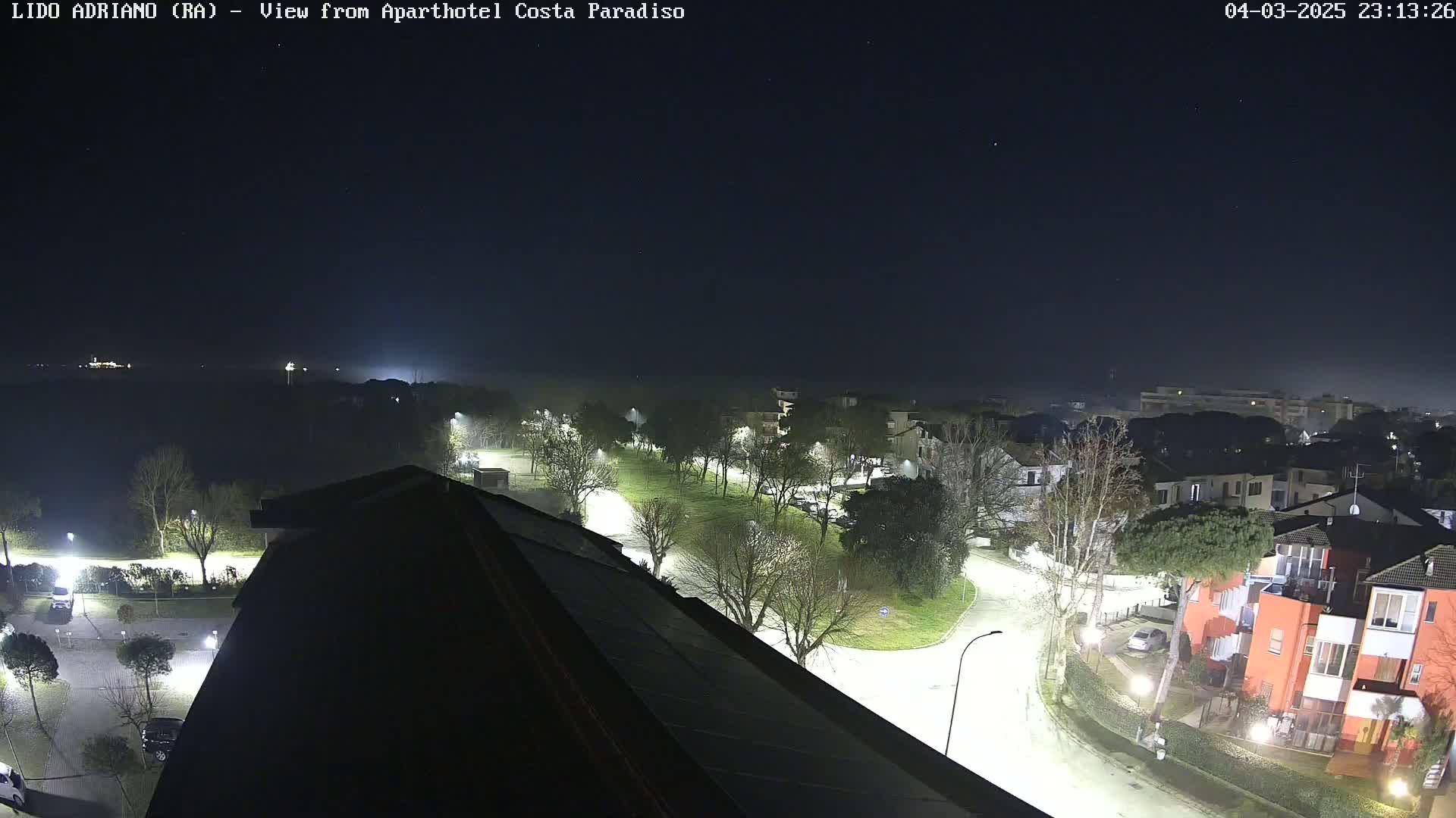 A nighttime, slightly hazy view of a residential area with well-lit streets and trees, seen from a rooftop.