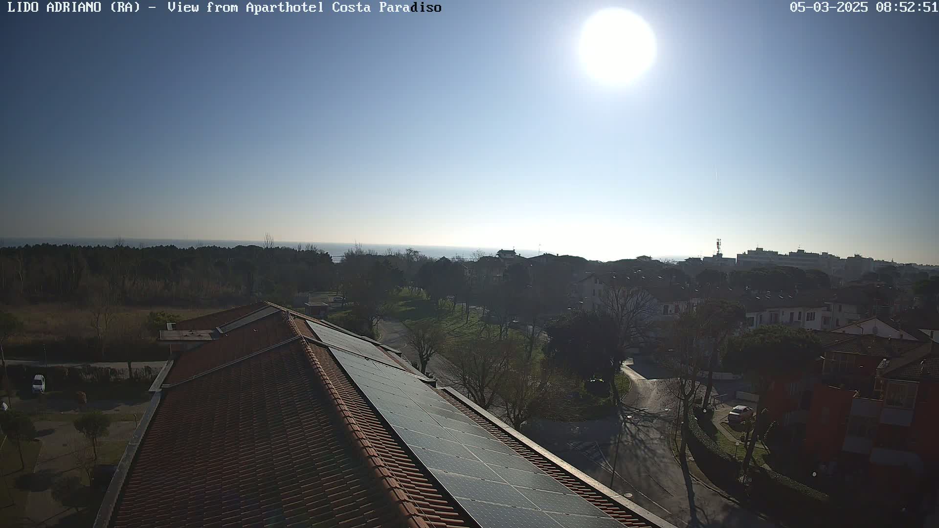 A sunny day reveals a coastal town viewed from a rooftop with solar panels, overlooking a tranquil sea.