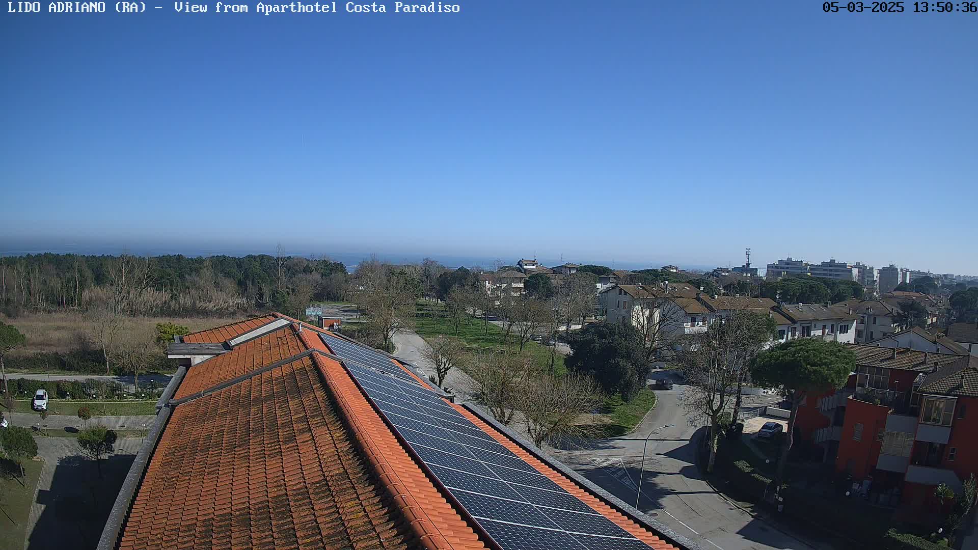 A rooftop with solar panels overlooks a residential area and a calm sea under a clear blue sky.
