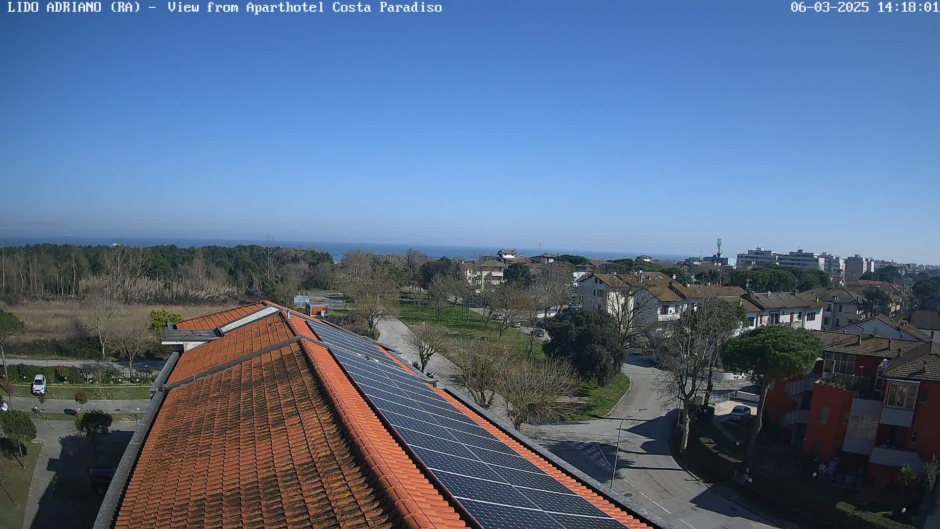 A rooftop with solar panels overlooks a residential area and a calm ocean under a clear blue sky.