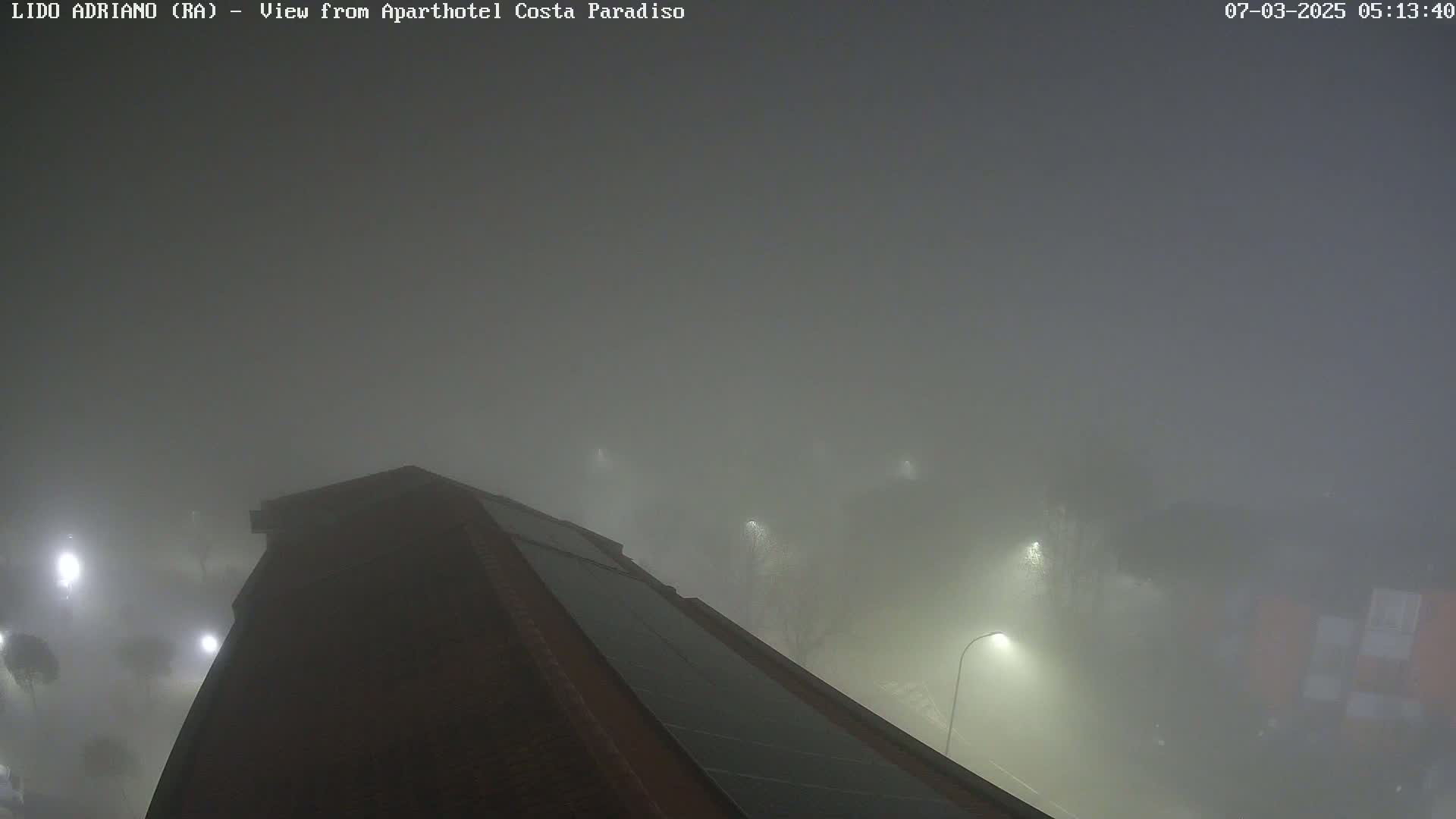 A dark building is partially visible in a dense fog at night, illuminated by several streetlights.