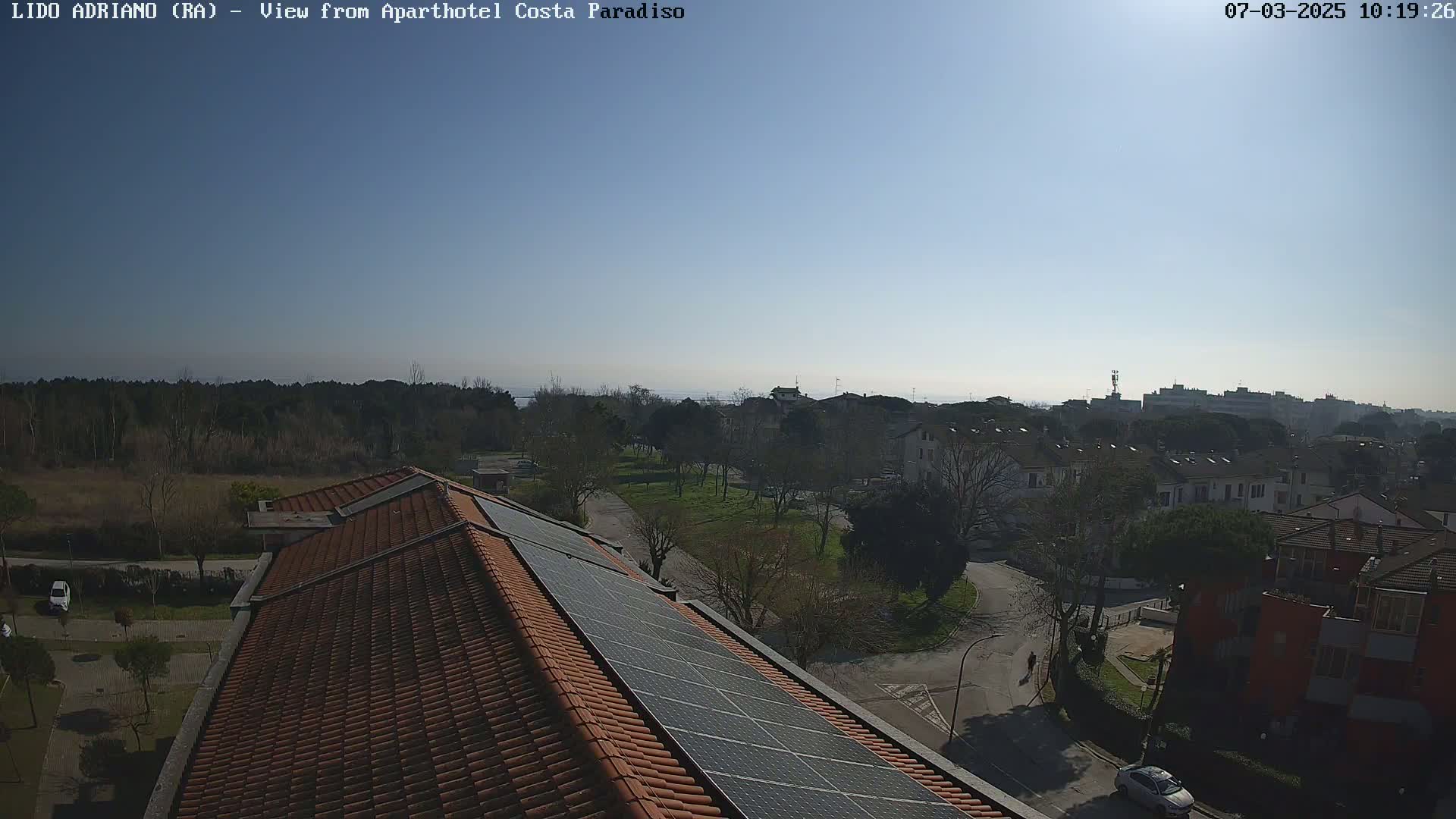 A high-angle, long shot on a clear, sunny day shows a rooftop with solar panels overlooking a residential area with trees and a distant body of water.