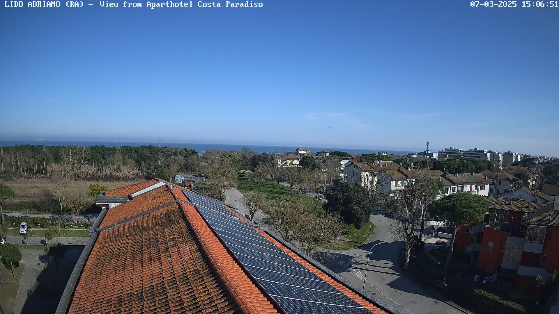 A rooftop with solar panels overlooks a residential area and a calm ocean under a clear blue sky.